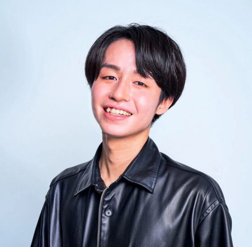 白い背景の前で黒いレザージャケットを着用し、明るく笑顔を見せる若いアジア人男性のポートレート写真です。