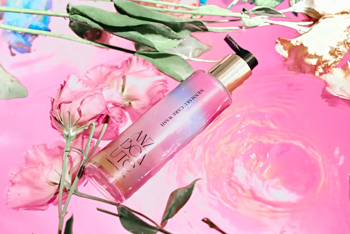 ピンク色の液体に浮かぶ、グラデーションが美しい化粧品のボトル。周りにはピンク色の花や緑の葉が添えられ、清潔感と華やかさを兼ね備えた美容製品のイメージを伝えています。