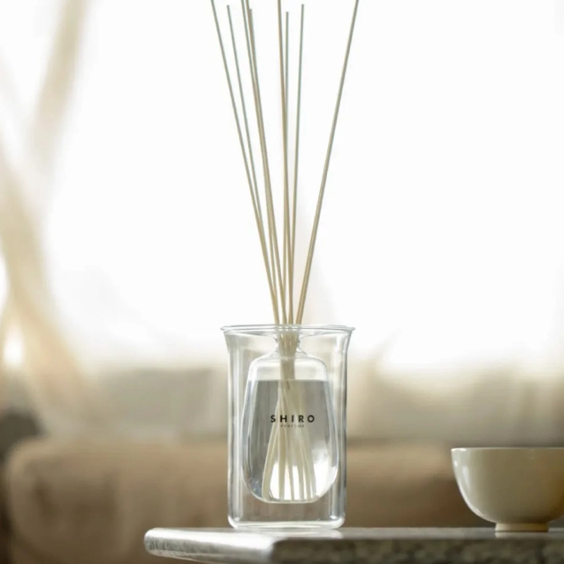明るい室内で、SHIROのロゴが入ったガラス製アロマディフューザーが置かれている様子を捉えた画像です。細いスティックが複数挿されており、その隣には白いカップが見えます。