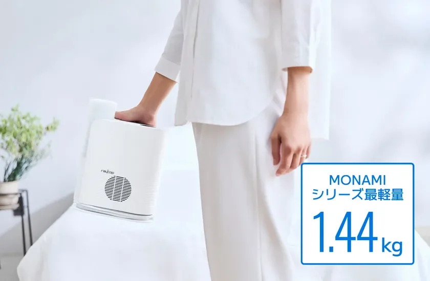 白い服を着た人物が、白い布団の上に置かれた「raycop MONAMI」という軽量の布団クリーナー（1.44kg）の横に立っている画像です。清潔感のある寝室の様子が伺えます。