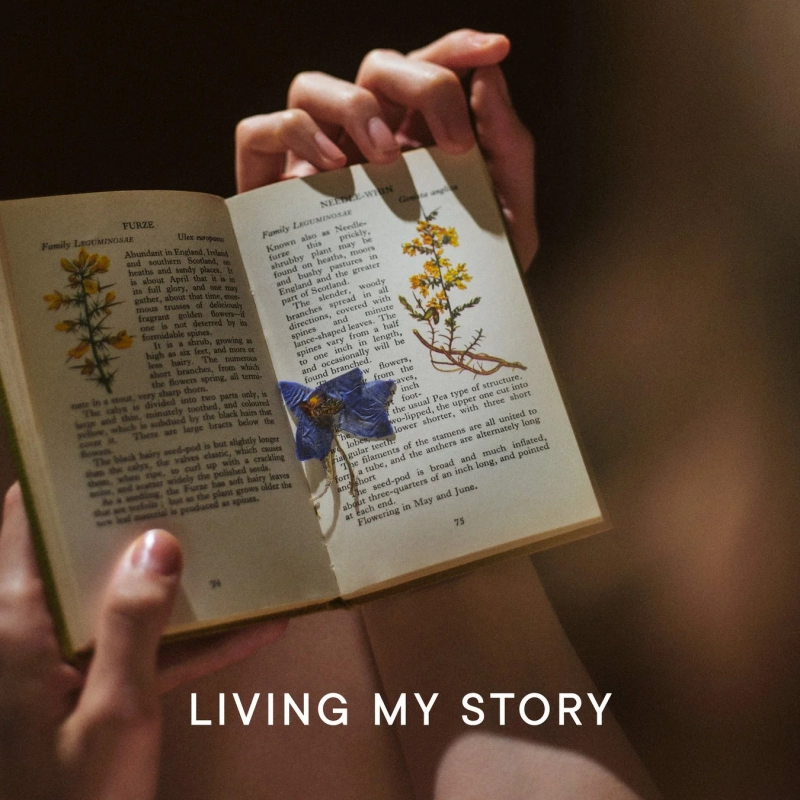 暗い背景の中、手が古い本を開いており、ページには植物のイラストと詳細な説明文が書かれています。青い押し花が挟まれ、「LIVING MY STORY」という文字が重ねられています。