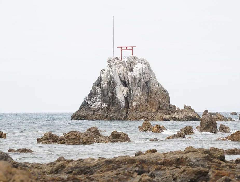 海にそびえ立つ大きな岩の上に赤い鳥居が立つ、日本の神秘的な海岸風景です。波打ち際には小さな岩々が点在し、静かで美しい自然の景色が広がっています。