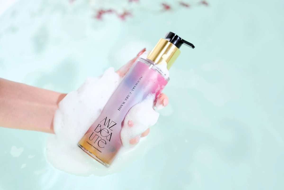 泡立つ水の中に手で持たれたピンクとゴールドのポンプ式ボトルが写っており、バスタイムでのスキンケアやリラックスを連想させる画像です。