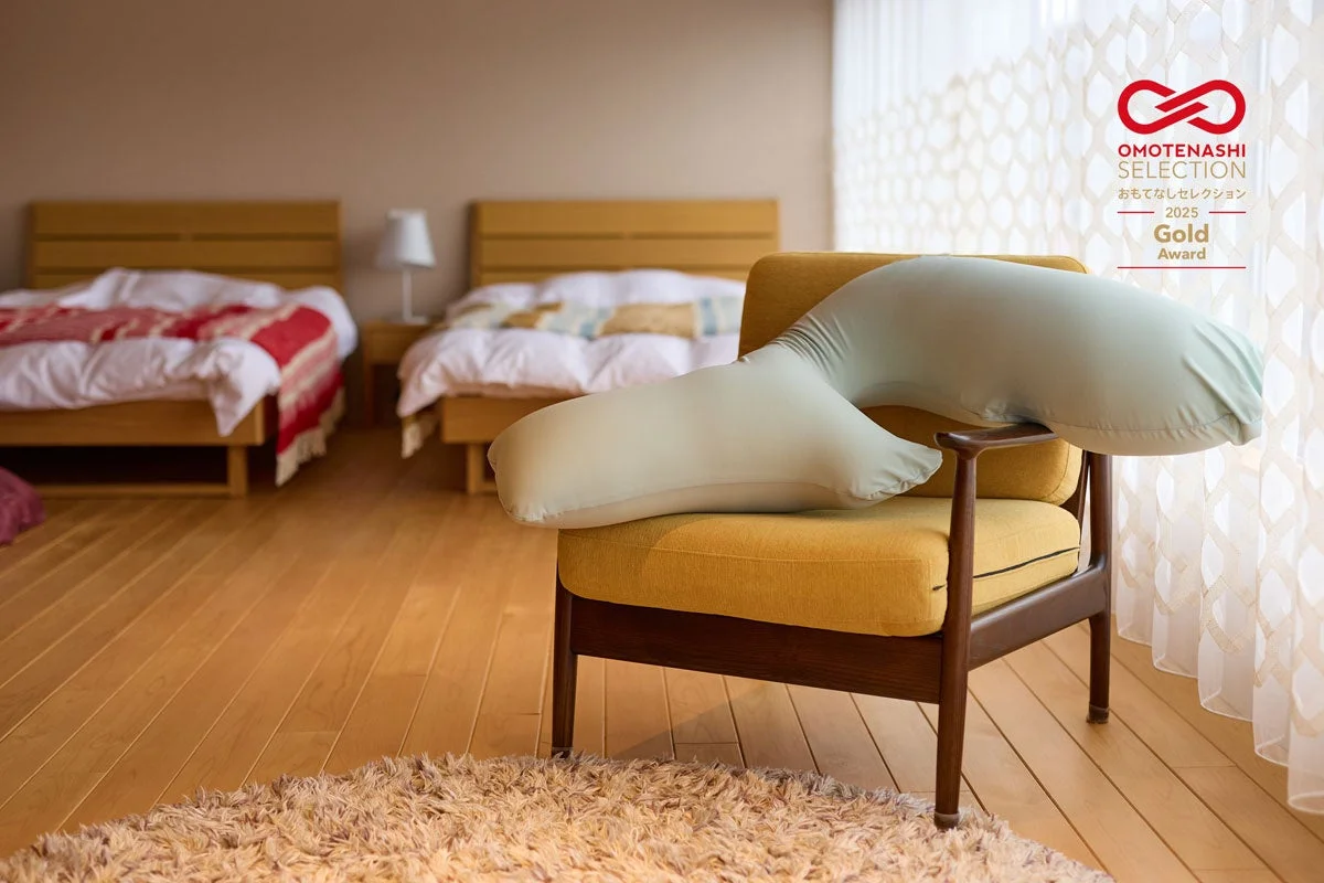 木製の床とベッドが特徴的な寝室の画像です。手前にはマスタード色のソファに抱き枕が置かれ、奥には2つの整えられたベッドが見えます。おもてなしセレクション2025ゴールドアワードのロゴがあり、快適な空間を演出しています。