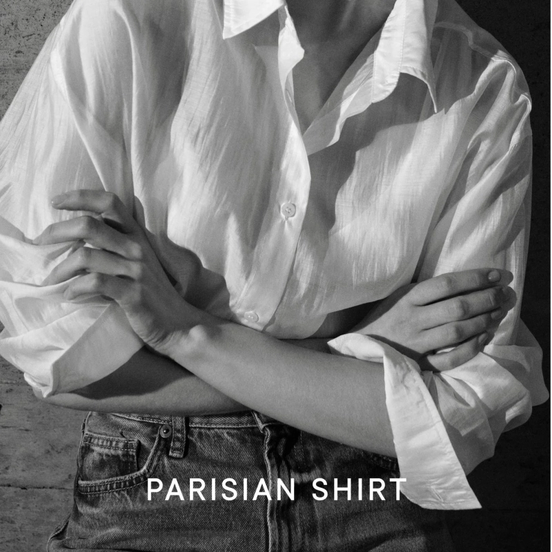 モノクロで撮影された、白いシャツとデニムを着用した人物のポートレート。腕を組んだポーズで、シャツの質感やカジュアルなスタイルが強調されています。テキスト「PARISIAN SHIRT」が表示されています。
