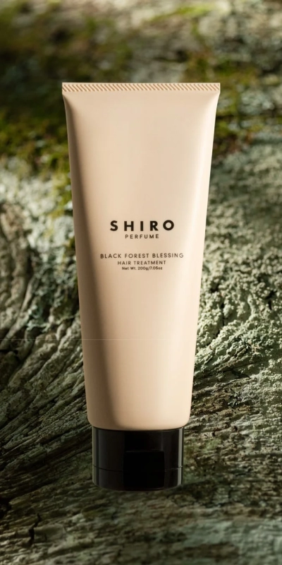 SHIROの「BLACK FOREST BLESSING」ヘアトリートメントのチューブが、苔むしたような自然な背景に置かれている製品写真です。ベージュ色の容器と黒いキャップが特徴的です。