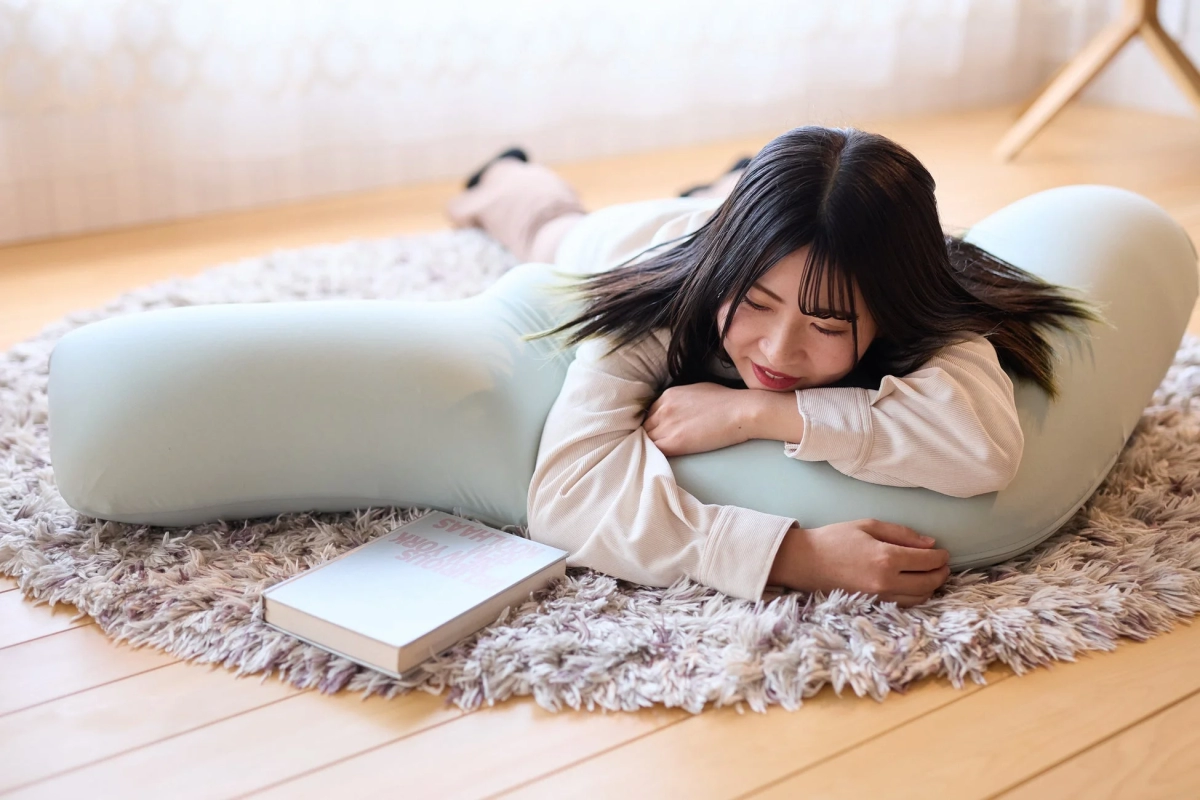 シャギーラグの上に置かれた淡い緑色の抱き枕にうつ伏せになり、リラックスした表情でくつろぐ若い女性。傍らには一冊の本が置かれている。