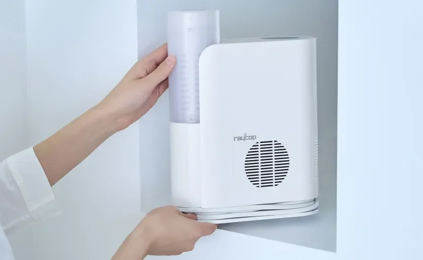 白い壁のくぼみに置かれた白い家電製品に、人が透明な水タンクをセットしている様子。raycopのロゴが見え、加湿器か空気清浄機のメンテナンス風景と推測されます。