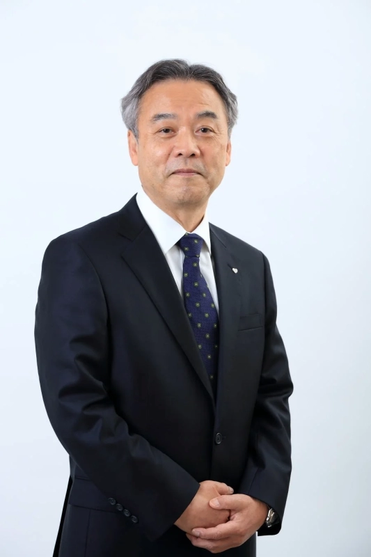 白い背景の前に立つ、スーツ姿の中年男性のポートレート。彼はカメラを真っ直ぐ見つめ、両手を前で組んでいます。落ち着いた表情をしており、ビジネスシーンや公式な場面に適した印象を与えます。